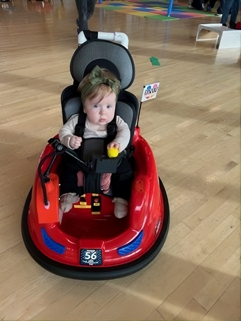 A baby with a green bow grins from the driver's seat of a red adapted Wayfinder™ ride-on car.